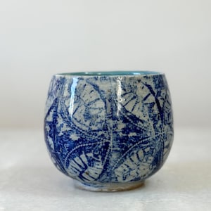 May include: A handmade ceramic cup with a rounded shape. The cup features a blue and white pattern on the exterior and a light blue interior. The design appears to be hand-painted, giving it a unique, artistic look. The cup is suitable for beverages.