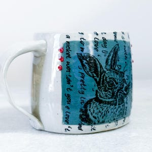 May include: A white ceramic mug featuring a teal rectangular panel with a black ink illustration of a rabbit. The design includes handwritten text and three small red dots. The mug has a curved handle and a smooth, glossy finish throughout.