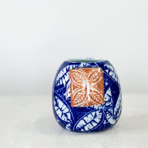 May include: A small, blue ceramic vase with a white leaf pattern and a square, orange floral design. The vase has a rounded shape and a small opening at the top. The background is white.