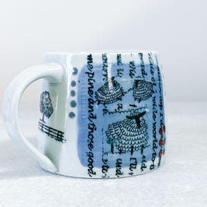 May include: A white ceramic mug featuring a blue rectangular patch with black line drawings of sheep and cursive text. The mug also displays a black illustration of a tree and a fence near the handle, along with small red dots and blue circles.