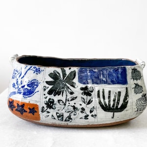 May include: A handcrafted ceramic bowl with a rectangular shape. The exterior features a patchwork design with blue, black, and orange floral and animal motifs. The interior is a deep blue color, and the bowl has two small handles.
