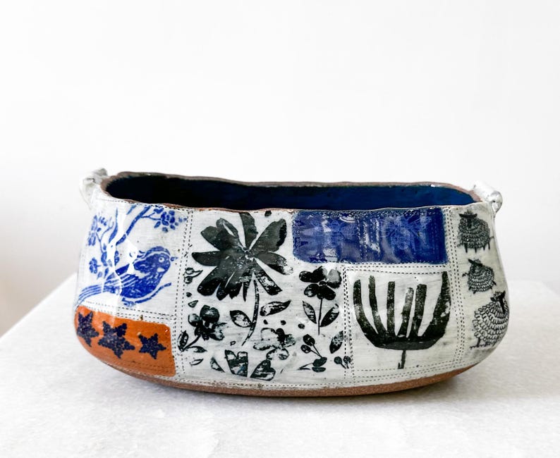 May include: A handcrafted ceramic bowl with a rectangular shape. The exterior features a patchwork design with blue, black, and orange floral and animal motifs. The interior is a deep blue color, and the bowl has two small handles.