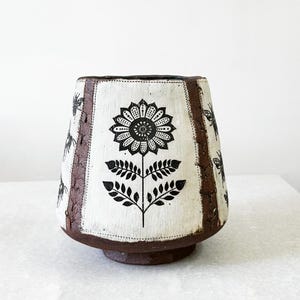 May include: A handcrafted ceramic vase with a wide, cylindrical shape. The vase features a cream-colored background with a large black sunflower design on the front. The sides have black insect illustrations and brown trim. The base is a dark brown.