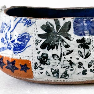May include: A handcrafted ceramic bowl with a patchwork design. The bowl features blue and black floral and bird motifs on a white background, with an orange and blue border. The interior is a deep blue color.