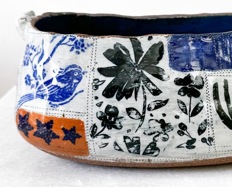 May include: A handcrafted ceramic bowl with a patchwork design. The bowl features blue and black floral and bird motifs on a white background, with an orange and blue border. The interior is a deep blue color.