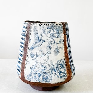 May include: A ceramic vase with a unique design. The vase features a blue and white floral pattern with a hummingbird, brown leather-like trim, and a brown base. The vase has a height of approximately 7 inches.