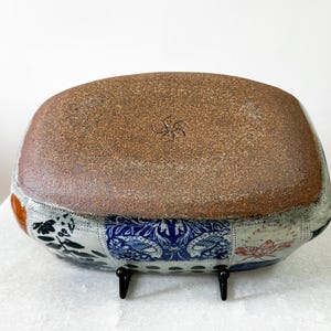 May include: A rectangular ceramic bowl with a brown, textured base and a patchwork design around the sides. The patchwork includes floral and geometric patterns in blue, white, and orange. The bowl is supported by two small black stands.
