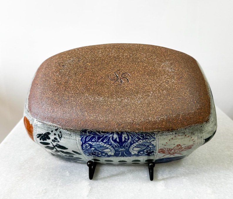 May include: A rectangular ceramic bowl with a brown, textured base and a patchwork design around the sides. The patchwork includes floral and geometric patterns in blue, white, and orange. The bowl is supported by two small black stands.