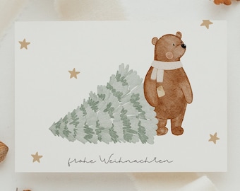 May include: A white card with a brown bear wearing a white scarf standing next to a green Christmas tree. The card has gold stars and the text "frohe Weihnachten" written in black.