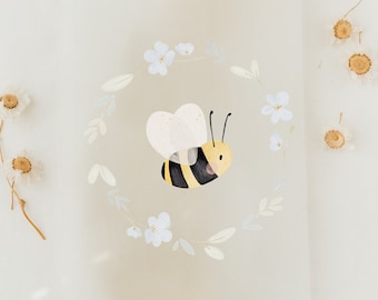 May include: A white card with a watercolor illustration of a bee flying in a circle of white flowers. The card is on a white background with a beige ribbon.