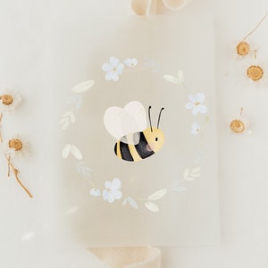 Op de afbeelding: Een witte kaart met een aquarel-illustratie van een bij die in een cirkel van witte bloemen vliegt. De kaart ligt op een witte achtergrond met een beige lint.