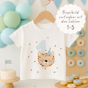 Könnte beinhalten: Weißes Kurzarm-T-Shirt mit einem Cartoon-Leoparden-Gesicht mit Partyhut und der Zahl 1 in Hellblau. Das T-Shirt hängt an einem Holzbügel. Der Text auf dem Bild lautet "Bügelbild verfügbar mit den Zahlen: 1-5".