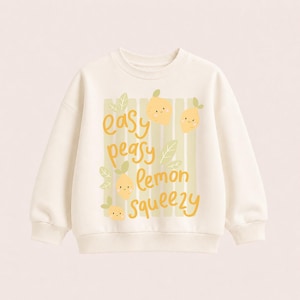Könnte beinhalten: Ein cremefarbenes Sweatshirt mit Rundhalsausschnitt und einem Grafikdruck von vier lächelnden gelben Zitronen mit grünen Blättern. Der Text "easy peasy lemon squeezy" ist in gelber Schreibschrift auf hellgrünen vertikalen Streifen gedruckt.