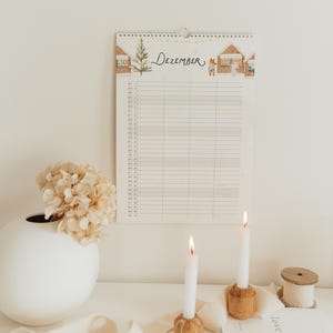May include: A wall calendar for the month of Dezember featuring a watercolor illustration of a Christmas tree and small houses with a fox and a ghost. The calendar has a grid layout for daily entries and is printed on white paper with a spiral binding.