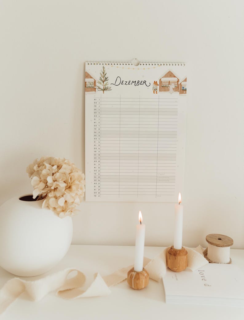 May include: A wall calendar for the month of Dezember featuring a watercolor illustration of a Christmas tree and small houses with a fox and a ghost. The calendar has a grid layout for daily entries and is printed on white paper with a spiral binding.