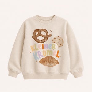 Könnte beinhalten: Ein beigefarbener Rundhals-Pullover mit einem Aquarell-Motiv, das eine Brezel, einen Keks mit Schokostückchen und ein Croissant zeigt, die jeweils ein kleines lächelndes Gesicht haben. Der Text "KLEINER KRÜMEL" ist in bunten, abgerundeten Buchstaben gedruckt.