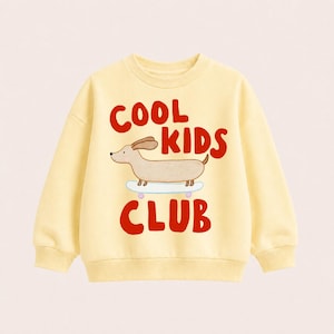 Könnte beinhalten: Ein hellgelbes Langarm-Sweatshirt mit einer grafischen Illustration eines beigen Dackels auf einem hellblauen Skateboard. Der Text "COOL KIDS CLUB" ist in fetten roten Buchstaben über und unter der Hundeillustration auf der Vorderseite gedruckt.