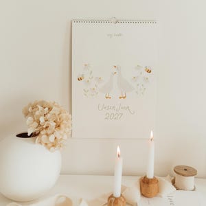 May include: A wall calendar titled Unser Jahr 2027 featuring a watercolor illustration of two white geese surrounded by daisies and bees. The text Hej Hanni is printed at the top. The calendar is white with a spiral binding at the top edge.