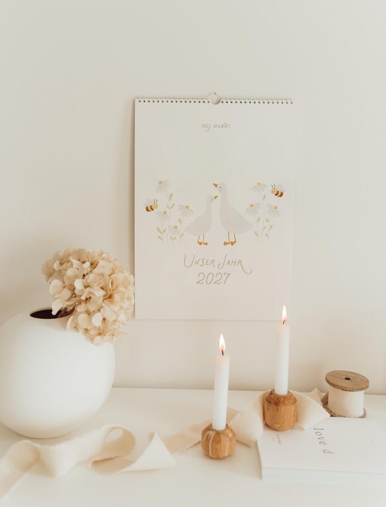 May include: A wall calendar titled Unser Jahr 2027 featuring a watercolor illustration of two white geese surrounded by daisies and bees. The text Hej Hanni is printed at the top. The calendar is white with a spiral binding at the top edge.