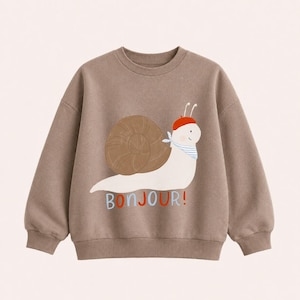 Könnte beinhalten: Ein taupefarbenes Langarm-Sweatshirt mit einer Grafik einer weißen Schnecke mit braunem Gehäuse, die ein rotes Barett und ein blau-weiß gestreiftes Halstuch trägt. Der Text BONJOUR! steht unter der Schnecke in blauen und roten Buchstaben.