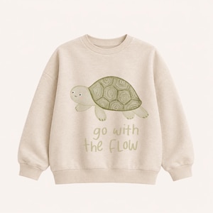 Könnte beinhalten: Ein beigefarbenes Sweatshirt mit Rundhalsausschnitt und einer Aquarell-Illustration einer hellgrünen Schildkröte. Unter der Schildkröte steht der Text "go with the FLOW" in einer passenden grünen Handschrift-Schriftart. Das Kleidungsstück hat lange Ärmel.