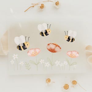 Könnte beinhalten: Aquarellillustration mit drei Bienen mit schwarz-gelben Streifen, die über rosa und orangefarbenen Blumen schweben. Kleine weiße Blumen und grüne Blätter befinden sich unten. Der Hintergrund ist eine weiche, neutrale Farbe.