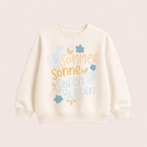 Könnte beinhalten: Ein cremefarbenes Langarm-Sweatshirt mit einem Grafikdruck, der den Text "SOMMER SONNE Seifen BLASEN" in Gelb und Blau zeigt. Das Design enthält Illustrationen von hellblauen Seifenblasen, kleinen blauen Blumen und grünen Blättern.