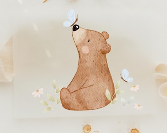 May include: A watercolor illustration of a brown bear sitting with its head tilted up, looking at a blue butterfly. The bear is surrounded by green leaves and white flowers.
