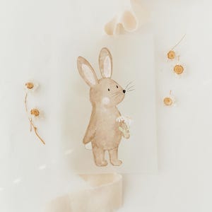 Op de afbeelding: Een aquarel-illustratie van een bruin konijn dat een boeket witte bloemen vasthoudt. Het konijn staat op een witte achtergrond met verspreide gedroogde bloemen.