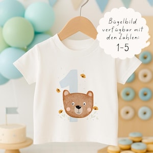 Könnte beinhalten: Ein weißes T-Shirt mit einer hellblauen Zahl 1 und einem braunen Bärengesicht. Der Bär hat eine schwarze Nase und kleine Augen. Das T-Shirt hängt an einem Holzbügel. Der Text auf dem Bild lautet "Bügelbild verfügbar mit den Zahlen: 1-5".