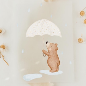 Op de afbeelding: Waterverfillustratie van een bruine beer die een witte paraplu met gouden stippen vasthoudt. De beer staat op een wolk, met regendruppels die vallen. Het kunstwerk heeft een zacht, pastelkleurig kleurenpalet.