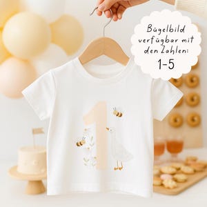 Op de afbeelding: Wit T-shirt met korte mouwen met een lichtperzik nummer 1, bijen en een ganzenafbeelding. Het shirt hangt aan een houten hanger. Tekst op de afbeelding luidt: "Strijkapplicatie beschikbaar met de nummers: 1-5."