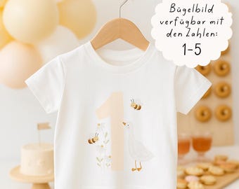 Puede incluir: Camiseta blanca de manga corta con un número 1 melocotón claro, abejas y un gráfico de ganso. La camiseta está colgada de una percha de madera. El texto de la imagen dice: "Imagen termoadhesiva disponible con los números: 1-5."