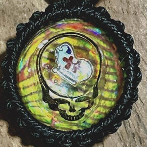 May include: A round pendant with a black macrame cord. The pendant features a skull design with a heart and cross symbol in the center. The background has a colorful, iridescent pattern.