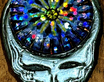 May include: A silver pendant featuring a stylized skull design. The pendant has a circular center with a colorful, glittery, abstract design in shades of blue, green, yellow, and red. The skull has a detailed, textured appearance.