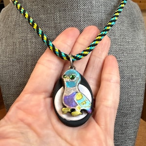 May include: A black and yellow braided cord necklace with a silver pendant featuring a blue, green, and purple bird on a white and black background.