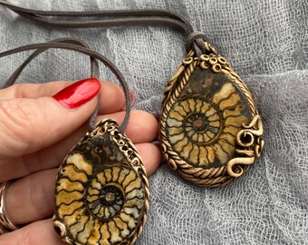 May include: Two gold-toned pendants with a brown and white ammonite fossil design. The pendants are on a gray suede cord.