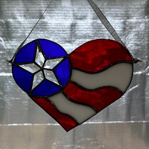May include: A heart-shaped stained glass piece featuring a patriotic design. The top left section is blue with a white star, while the rest of the heart displays red and white stripes. A black ribbon is attached for hanging.