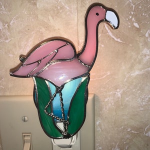May include: A pink flamingo nightlight with green leaves and a blue background. The nightlight is made of stained glass and has a white plug.