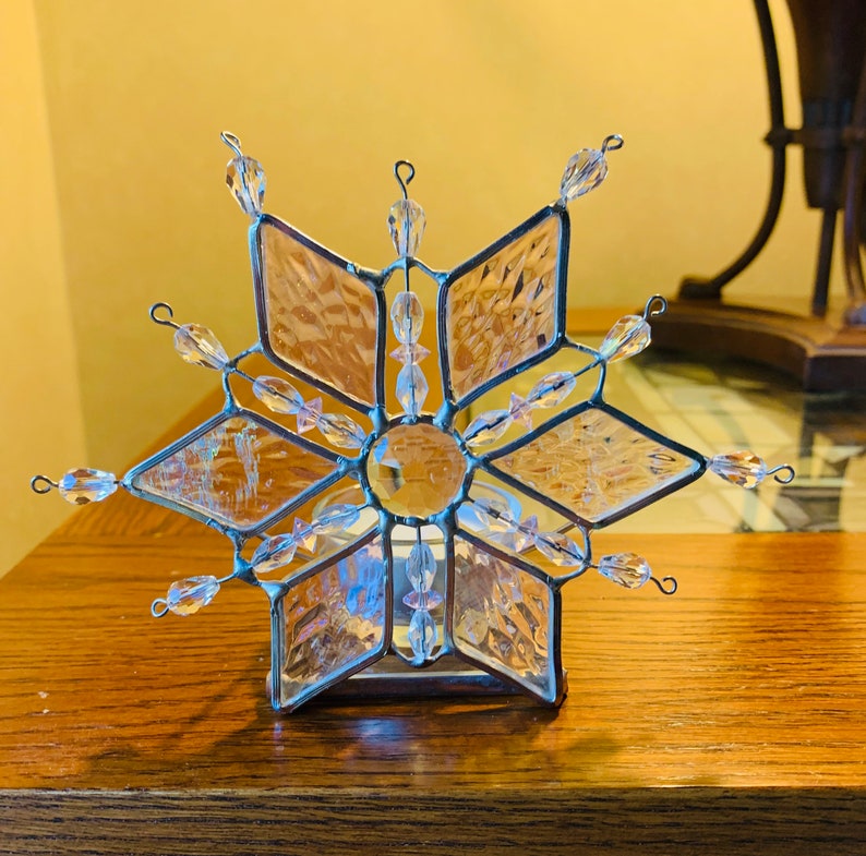 May include: A clear glass snowflake candle holder with silver accents and iridescent beads. The snowflake is a stained glass design with a clear glass center.