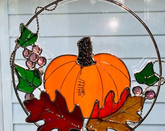 May include: A stained glass pumpkin wreath with green leaves, pink berries, and orange, red, and yellow fall leaves. The wreath is hanging on a window.