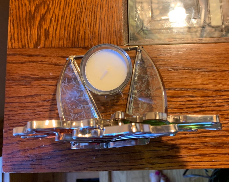 May include: A silver candle holder with a clear glass insert for a tea light candle. The holder is shaped like a sailboat with a green and orange sail.