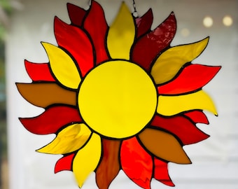 Könnte beinhalten: Eine Sonne aus Buntglas mit roten, orangen und gelben Glasscheiben. Die Sonne ist von einem Ring aus braunem Glas umgeben.
