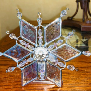 May include: A clear glass snowflake nightlight with pink beads and a crystal center. The snowflake is on a wooden surface.