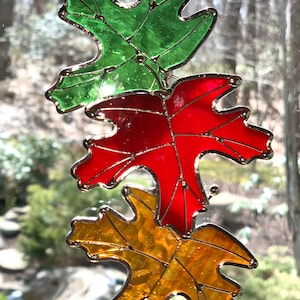May include: Three stained glass leaves hanging from a window. The top leaf is green, the middle leaf is red, and the bottom leaf is yellow.