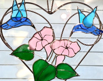 Pode incluir: Um apanhador de sol em forma de coração de vidro colorido com dois beija-flores azuis e flores de glória-da-manhã rosa. O apanhador de sol é feito de vidro e metal e é projetado para pendurar em uma janela.