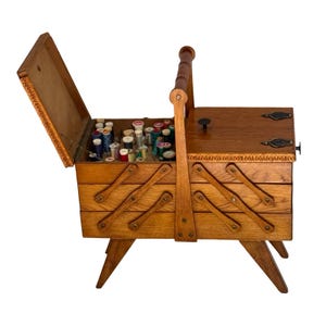 May include: A wooden accordion-style sewing box with a medium brown finish, featuring a central handle, four angled legs, and multiple tiered compartments. The top lid is open, revealing an interior filled with various colorful thread spools.