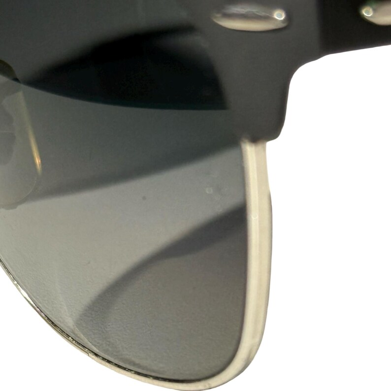 May include: Close-up of sunglasses with a black frame and grey lenses. The lenses are slightly curved, and the frame has silver accents. The sunglasses have a classic, timeless design.