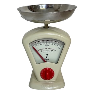 May include: A white kitchen scale with a silver bowl. The scale has a red knob and a dial with black markings. The dial reads "NOT LEGAL FOR USE IN TRADE" and "CAPACITY 1kg SENSITIVITY 100g". The scale is made by Dutton.
