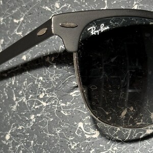 May include: Black Ray-Ban sunglasses with a matte finish. The sunglasses have a classic design with a browline and dark lenses. The Ray-Ban logo is visible on the lens. The frame is a dark grey colour.